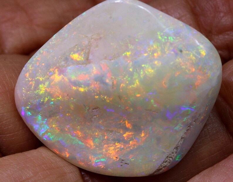 Może przedstawiać: Wypolerowany, biały opal z żywymi, iryzującymi błyskami koloru. Kamień szlachetny prezentuje grę światła z odcieniami pomarańczowego, ż&oacute;łtego, zielonego i niebieskiego. Opal jest gładki i zaokrąglony, ukazując swoje naturalne piękno.