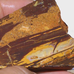 Puede incluir: Una piedra natural rugosa con una mezcla de colores. La piedra presenta capas de marr&oacute;n oscuro, beige y amarillo. La superficie tiene una apariencia texturizada con grietas y variaciones de color visibles. La piedra se sostiene en una mano.