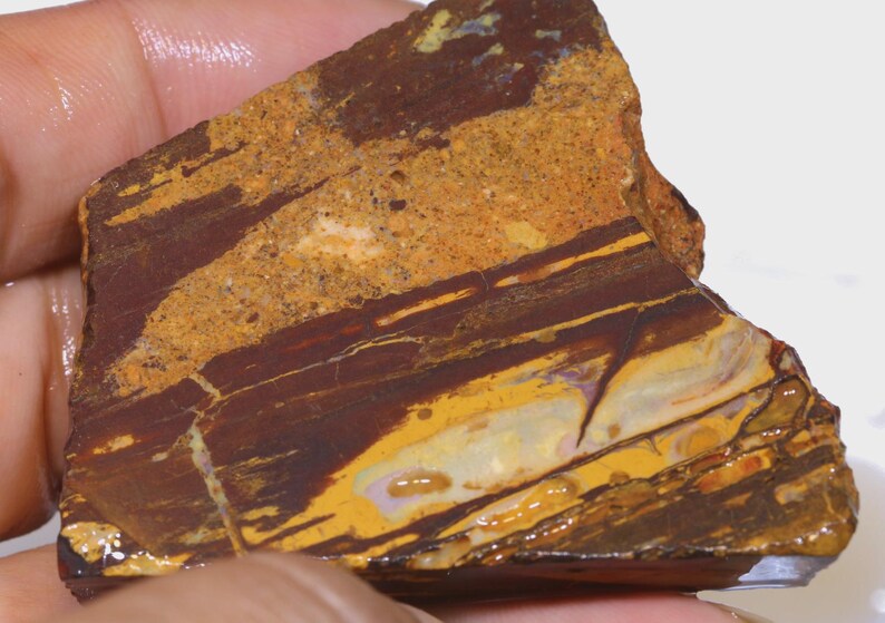 Puede incluir: Una piedra natural rugosa con una mezcla de colores. La piedra presenta capas de marr&oacute;n oscuro, beige y amarillo. La superficie tiene una apariencia texturizada con grietas y variaciones de color visibles. La piedra se sostiene en una mano.