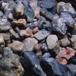 May include: A collection of raw, unpolished blue opal stones featuring irregular shapes and smooth, glossy surfaces. The stones display varying shades of greyish-blue, with some pieces showing small patches of white, orange, and iridescent color.