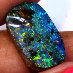 May include: A polished, rectangular opal gemstone with a vibrant display of color. The stone features a mix of blues, greens, and yellows, with dark inclusions. The opal's surface is smooth and reflective, showcasing its natural beauty.