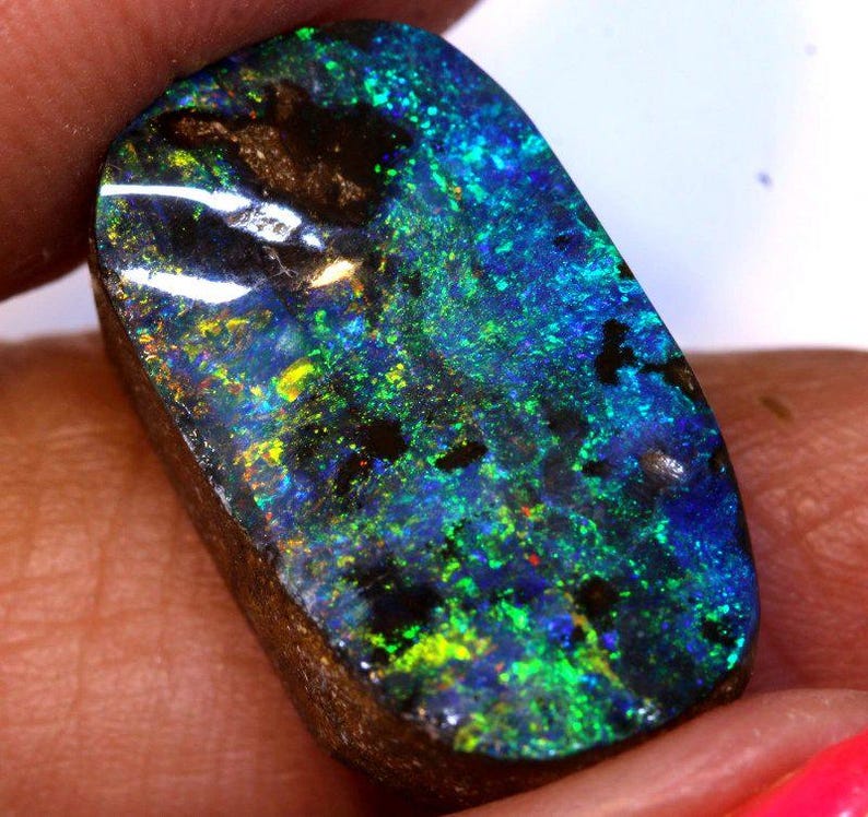 May include: A polished, rectangular opal gemstone with a vibrant display of color. The stone features a mix of blues, greens, and yellows, with dark inclusions. The opal's surface is smooth and reflective, showcasing its natural beauty.