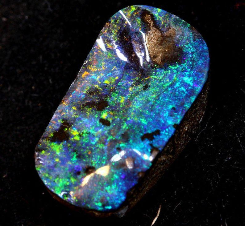May include: A polished black opal gemstone with vibrant blue, green, and yellow flashes. The opal has a dark base color with a play of color across its surface. The stone is cut into a rounded rectangular shape.