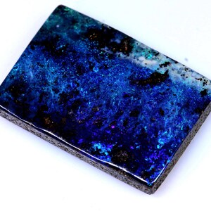May include: A rectangular gemstone displaying a rich palette of blues, greens, and blacks. The stone's surface has a textured appearance, with areas of deep colour and lighter, iridescent patches. The edges are a dark, contrasting colour.