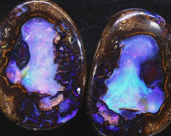 May include: Two polished opal gemstones with a dark brown matrix and vibrant internal colors. The opals display a play of color with blues, purples, and hints of green. The gemstones have an oval shape and are set against a black background.