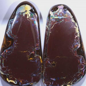 Könnte beinhalten: Zwei polierte, tropfenförmige Opale. Die Opale haben eine dunkelbraune Grundfarbe mit lebendigen Blitzen in Gelb, Blau und Grün. Die inneren Muster ähneln Blitzen oder Adern und erzeugen ein einzigartiges und auffälliges Aussehen. Die Opale sind vor einem weißen Hintergrund.