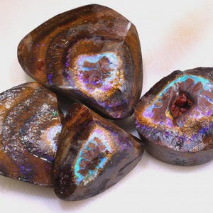 Puede incluir: Cuatro piedras de ópalo natural pulidas. Los ópalos tienen un color base marrón oscuro con destellos iridiscentes de azul, púrpura y dorado. Cada piedra tiene una forma y un patrón interno únicos, mostrando la belleza natural de la gema.