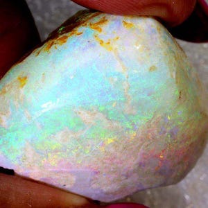 Puede incluir: Primer plano de una piedra preciosa de ópalo en bruto. La piedra muestra una mezcla de colores, incluyendo azul, verde, amarillo y rosa, con una base blanco lechoso. El ópalo está sostenido por los dedos de una persona.
