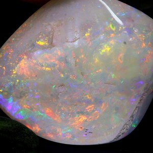 Może przedstawiać: Wypolerowany kamień szlachetny opalu o mlecznobiałej barwie. Opal wykazuje żywe błyski kolor&oacute;w, w tym pomarańczowy, ż&oacute;łty, zielony i fioletowy, na swojej powierzchni. Kamień ma gładki, zaokrąglony kształt i wysoki połysk.