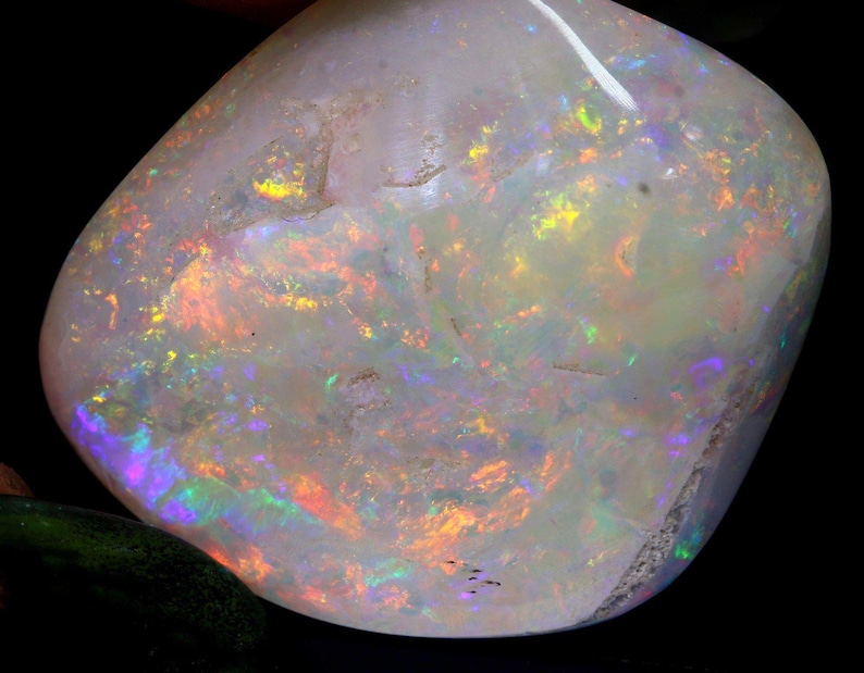 Może przedstawiać: Wypolerowany kamień szlachetny opalu o mlecznobiałej barwie. Opal wykazuje żywe błyski kolor&oacute;w, w tym pomarańczowy, ż&oacute;łty, zielony i fioletowy, na swojej powierzchni. Kamień ma gładki, zaokrąglony kształt i wysoki połysk.