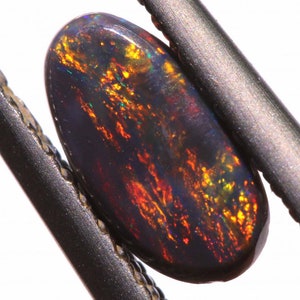Puede incluir: Una piedra preciosa de ópalo negro con un brillo iridiscente rojo, naranja y verde. La piedra de forma ovalada está sujeta por un par de pinzas metálicas.
