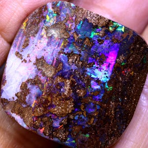 May include: A polished boulder opal gemstone with a square shape and rounded corners. The surface displays a vibrant play of colour with flashes of neon blue, purple, green, and red, interspersed with patches of textured, dark brown ironstone matrix.