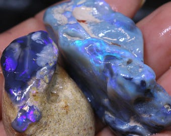 May include: Two rough, natural opal stones. One is a dark blue with purple flashes, the other is a light blue with green flashes.