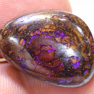 May include: A polished boulder opal gemstone featuring a dark brown and black ironstone matrix with vibrant, iridescent flashes of electric purple and deep red color play throughout the smooth, rounded, and organic surface of the stone.