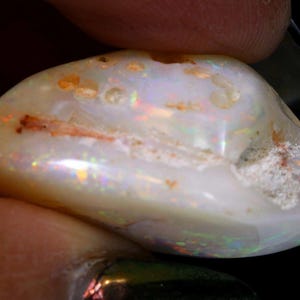 Może przedstawiać: Polerowany opal o owalnym kształcie, z kremowo-białą podstawą. Kamień szlachetny wykazuje iryzujące błyski kolor&oacute;w, w tym czerwony, zielony i ż&oacute;łty. Opal ma lekko szorstką teksturę z małymi, naturalnymi niedoskonałościami.