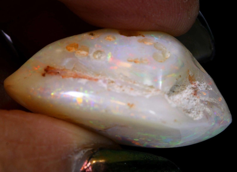 Może przedstawiać: Polerowany opal o owalnym kształcie, z kremowo-białą podstawą. Kamień szlachetny wykazuje iryzujące błyski kolor&oacute;w, w tym czerwony, zielony i ż&oacute;łty. Opal ma lekko szorstką teksturę z małymi, naturalnymi niedoskonałościami.