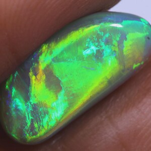 May include: A polished, oval-shaped opal gemstone. The opal displays a vibrant play-of-color, with flashes of green, yellow, and blue. The stone has a smooth, glossy surface and a translucent appearance, showcasing its internal structure and color variations.