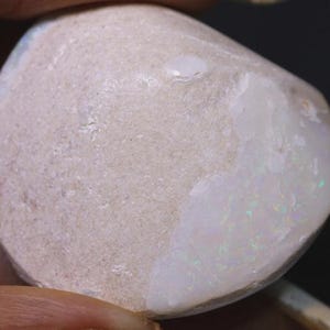 May include: A close-up of a polished opal gemstone. The stone has a rounded shape with a light beige, textured surface. One side of the opal displays a milky white color with iridescent flashes of green and blue.