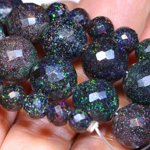 Puede incluir: Un collar de cuentas de ópalo negro facetadas con motas iridiscentes. Las cuentas, de diferentes tamaños, están ensartadas en un fino cordón transparente. Los ópalos muestran destellos verdes, azules y rojos.