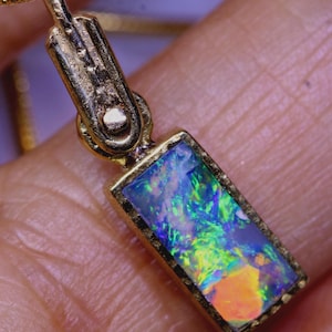 Könnte beinhalten: Ein goldener Anhänger mit einem rechteckigen Opal-Edelstein. Der Opal zeigt ein lebendiges Farbspiel mit Blau-, Grün- und Orangetönen. Der Anhänger hängt an einer zarten Goldkette und bildet ein elegantes Schmuckstück.