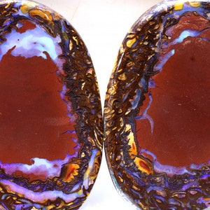 Könnte beinhalten: Zwei runde, polierte Opal-Cabochons mit einer dunkelroten Basis und einem blauen und violetten, irisierenden Rand. Die Opale haben ein natürliches, organisches Muster.