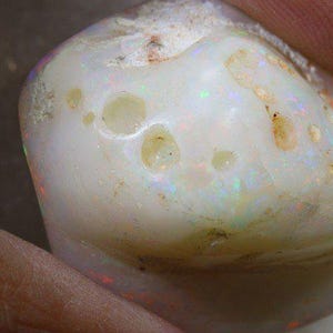 Może przedstawiać: Zbliżenie wypolerowanego kamienia szlachetnego opalu. Kamień jest gł&oacute;wnie biały z obszarami opalizującego koloru i małymi, nieregularnymi otworami. Powierzchnia ma gładki, zaokrąglony kształt z nutami brązu i ż&oacute;łci.