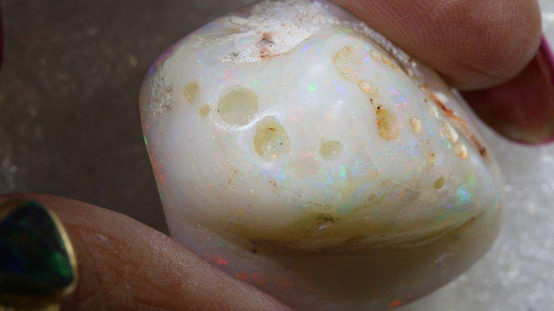Może przedstawiać: Zbliżenie wypolerowanego kamienia szlachetnego opalu. Kamień jest gł&oacute;wnie biały z obszarami opalizującego koloru i małymi, nieregularnymi otworami. Powierzchnia ma gładki, zaokrąglony kształt z nutami brązu i ż&oacute;łci.