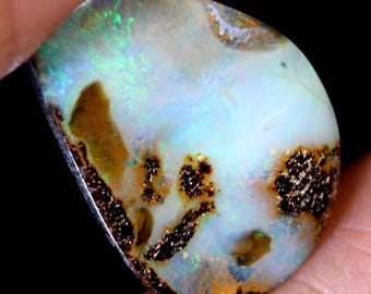 Puede incluir: Una piedra preciosa de ópalo en forma de lágrima con un cuerpo blanco e inclusiones de piedra de hierro marrón. El ópalo muestra un arcoíris de colores, incluyendo verde, azul y rojo.
