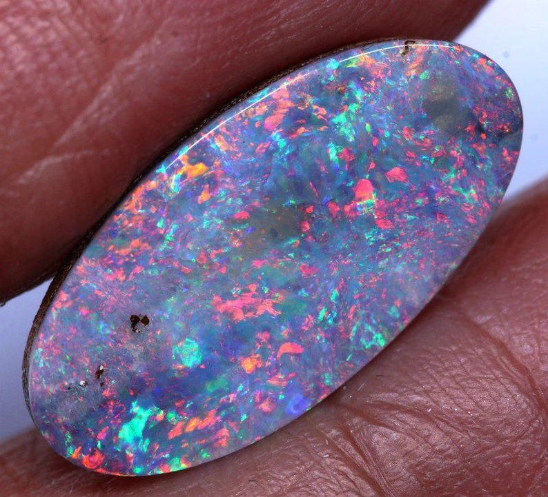 May include: An oval-shaped opal gemstone with a dark base color. The opal displays a vibrant play-of-color, with flashes of red, green, blue, and yellow throughout its surface. The gemstone is held between fingers, showcasing its polished finish.