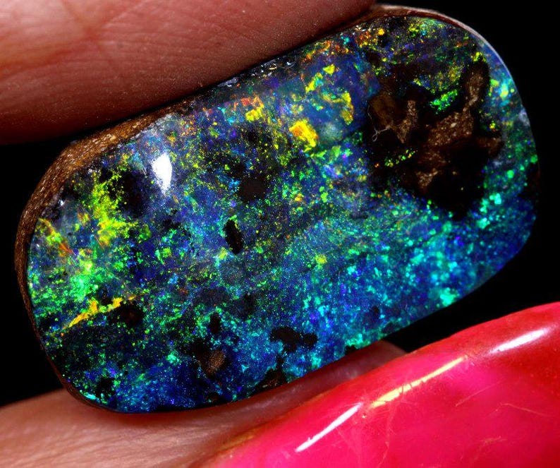 May include: A polished black opal gemstone with vibrant blue, green, and yellow flashes. The opal has a dark matrix with a natural, irregular shape. The gemstone is held between two fingers, showcasing its iridescent play-of-color.