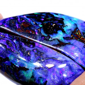 Könnte beinhalten: Ein großer, perlmuttartiger blauer Opal mit einem dunklen, irisierenden Schimmer. Der Opal ist in Tropfenform geschliffen und hat eine glatte, polierte Oberfläche.