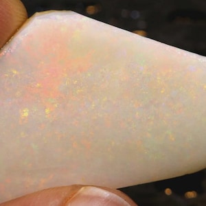 May include: A polished, irregularly shaped white opal gemstone featuring a translucent base with internal flashes of orange, yellow, and green light. The stone has a smooth, flat surface and exhibits a distinct play-of-color across its body.