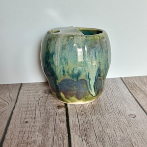 May include: A green and blue ceramic toothbrush holder with a unique glaze pattern. The holder has a rounded shape and a small opening for toothbrushes.