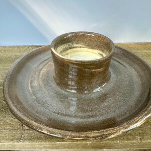 Puede incluir: Un plato de cerámica hecho a mano con un cuenco central elevado y una base circular ancha. El plato tiene una estética rústica y terrosa con un esmalte marrón moteado y un interior claro. Adecuado para servir salsas, aperitivos o platos pequeños.