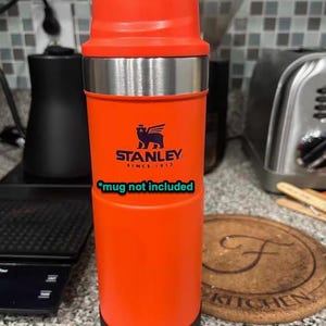 May include: Orange Stanley travel mug with a stainless steel lid and a black base. The mug has a logo that says "Stanley Since 1913" and the text "*mug not included".