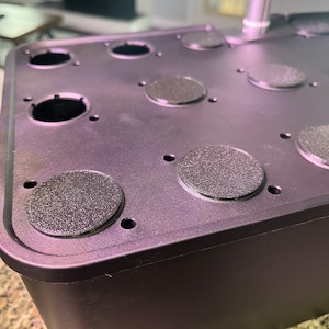 May include: Black plastic container with multiple round holes and black rubber plugs. The container is designed for growing plants.