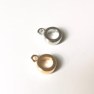 Könnte beinhalten: Zwei kleine, runde Schmuckkomponenten aus Metall mit einer Schlaufe oben. Eines ist silberfarben und das andere goldfarben. Die Komponenten sind vor einem weißen Hintergrund platziert.