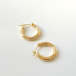 Peut inclure: Deux boucles d'oreilles créoles dorées. Les boucles d'oreilles sont rondes avec un design épais et lisse. Les boucles d'oreilles sont présentées sur une surface blanche. Ce sont des bijoux classiques.