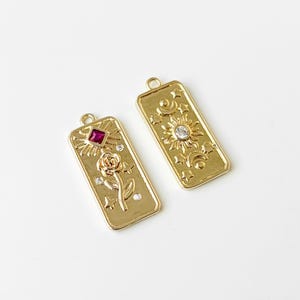 May include: Two gold-coloured rectangular pendants. One features a rose, a red square gem, and small clear gems. The other has a sun, moon, and stars with a clear gem. Both have a loop at the top for hanging.