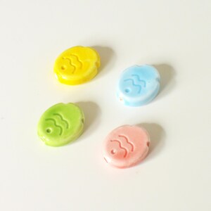 May include: Four oval-shaped ceramic beads in pastel yellow, light blue, lime green, and soft pink. Each bead features a small hole for threading and a debossed wavy line pattern on the surface, resembling a stylized fish or abstract design.