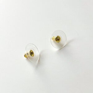 K&ouml;nnte beinhalten: Zwei transparente Ohrstecker aus Kunststoff mit goldenen Metallstiften. Die R&auml;nder sind rund und haben eine glatte Oberfl&auml;che.