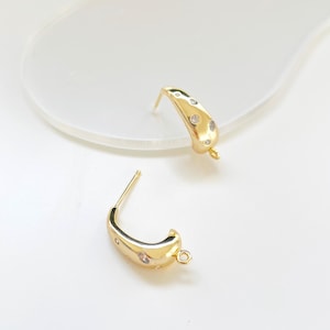 May include: A pair of gold-colored earrings with a curved design. Each earring features three small, clear gemstones and a loop at the bottom. The earrings are set against a white background.