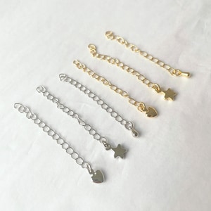 May include: Four silver and gold chain extenders with star and heart charms. The extenders are different lengths and are arranged in a row on a white background.
