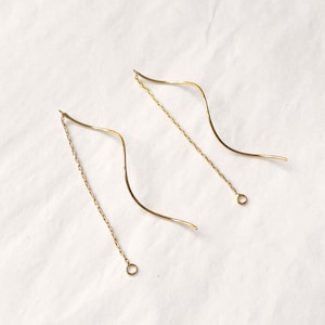 Könnte beinhalten: Ein Paar goldfarbene Ohrringe mit Faden. Jeder Ohrring hat ein geschwungenes, wellenartiges Design mit einer zarten Kette, die vom Ohr herabhängt. Die Ohrringe werden auf einer weißen Oberfläche präsentiert.