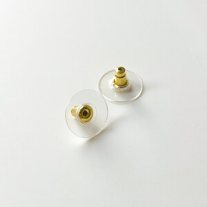 K&ouml;nnte beinhalten: Zwei transparente Ohrstecker aus Kunststoff mit goldenen Metallstiften. Die R&auml;nder sind rund und haben ein kleines Loch in der Mitte.