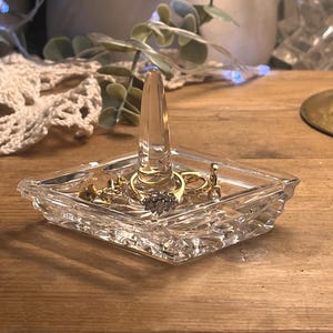 May include: A clear, square glass ring holder with a central cone for rings. Several gold and silver rings are displayed within. The holder is on a wooden surface.