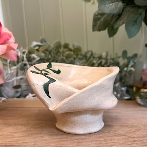 May include: A handcrafted, cream-coloured ceramic bowl with a distinctive, twisted form. The bowl has a speckled finish and is decorated with dark green, abstract leaf-like designs. The item is set against a blurred backdrop with floral elements.