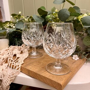 May include: Two crystal brandy glasses with diamond cut patterns. The glasses have a rounded bowl and a stemmed base. They sit on a wooden board, which rests on a white surface. Greenery is in the background.