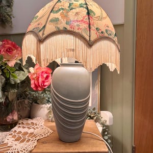 May include: A vintage table lamp featuring a floral lampshade with a fringed edge. The base is a light grey colour with a textured design. The lamp sits on a wooden table, alongside a lace doily.