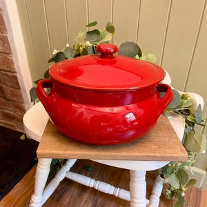May include: A bright red ceramic cooking pot with a matching lid and two side handles. The pot has a smooth, glossy finish and a rounded, bulbous shape. The lid features a small, centred knob handle on top, completing the classic kitchen vessel.